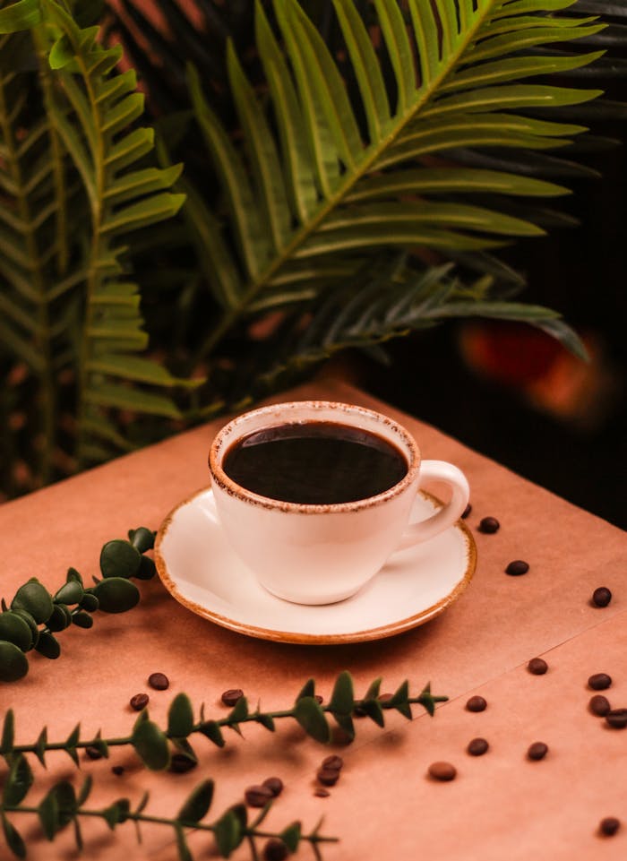 Aesthetic coffee scene with a white cup on a table, surrounded by lush green plants, and coffee beans scattered around.